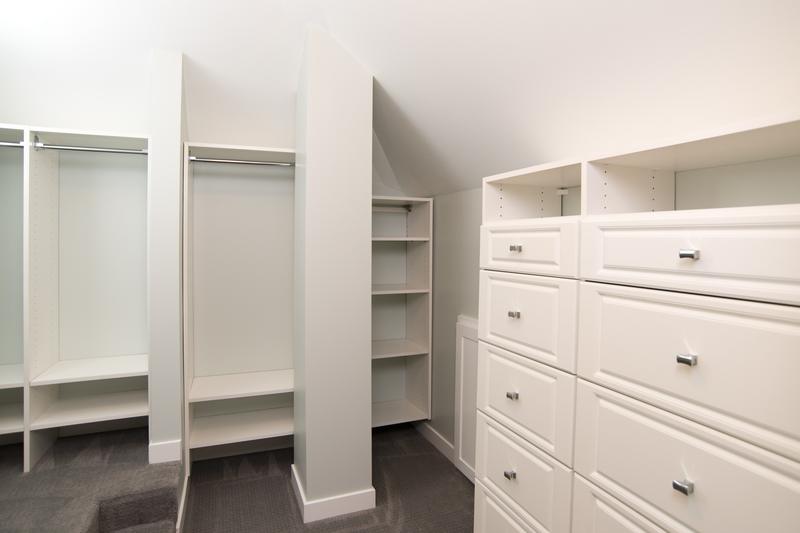 Walk-in closet with white built-in drawers, shelves and hanging rods under a sloped ceiling